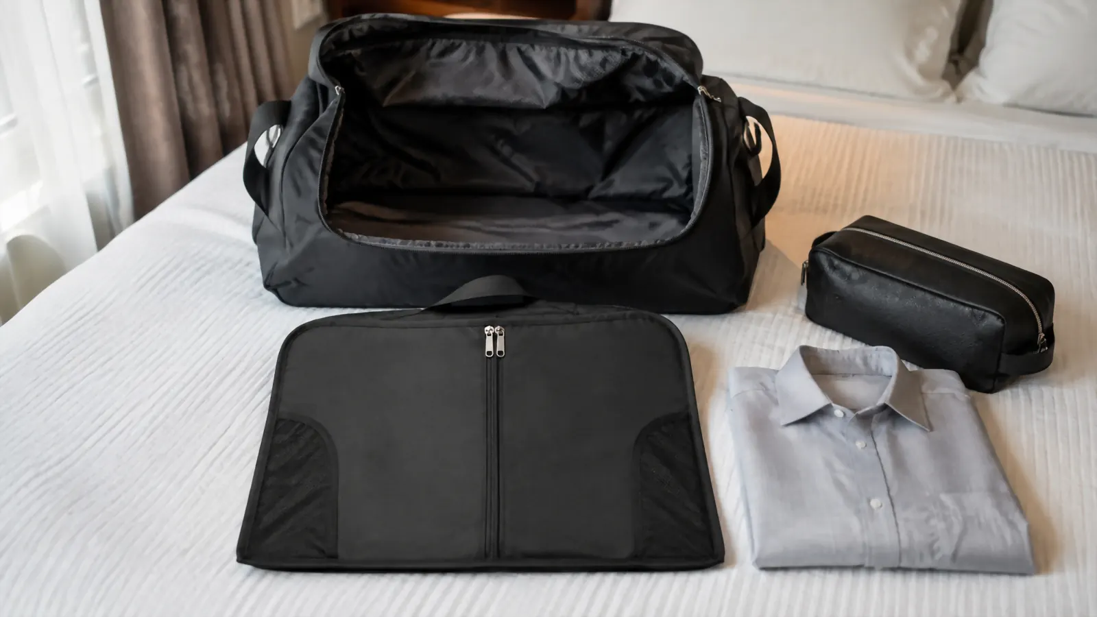 Folded garment bag placed flat along the bottom of an open duffel bag before the rest of the packing goes on top