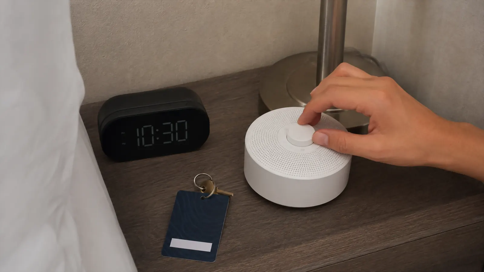 A hand adjusting the dial on a small white sound machine sitting on a hotel nightstand next to a lamp and a room key