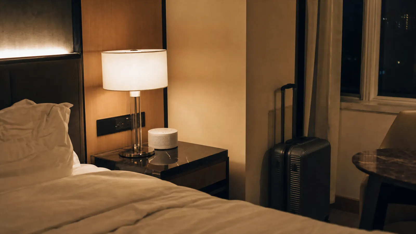 A dim hotel room at night with a small white noise machine glowing softly on the nightstand beside a made bed