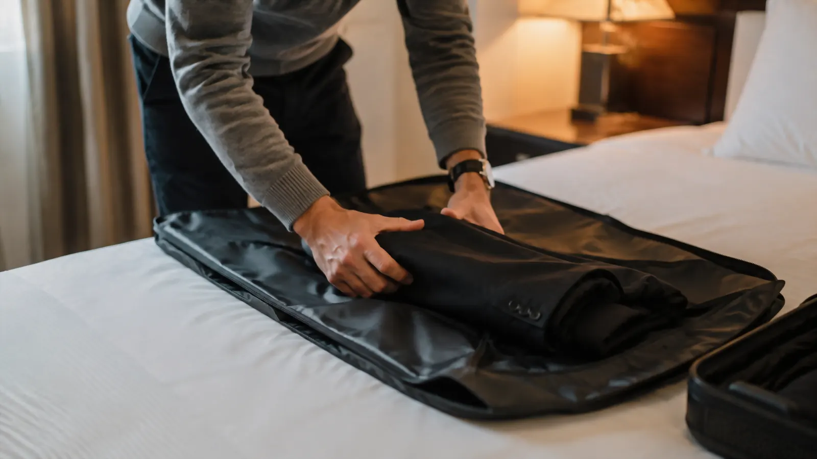 Traveler mid-fold, adjusting a slightly rumpled travel garment bag on a hotel bed during a first attempt at packing it
