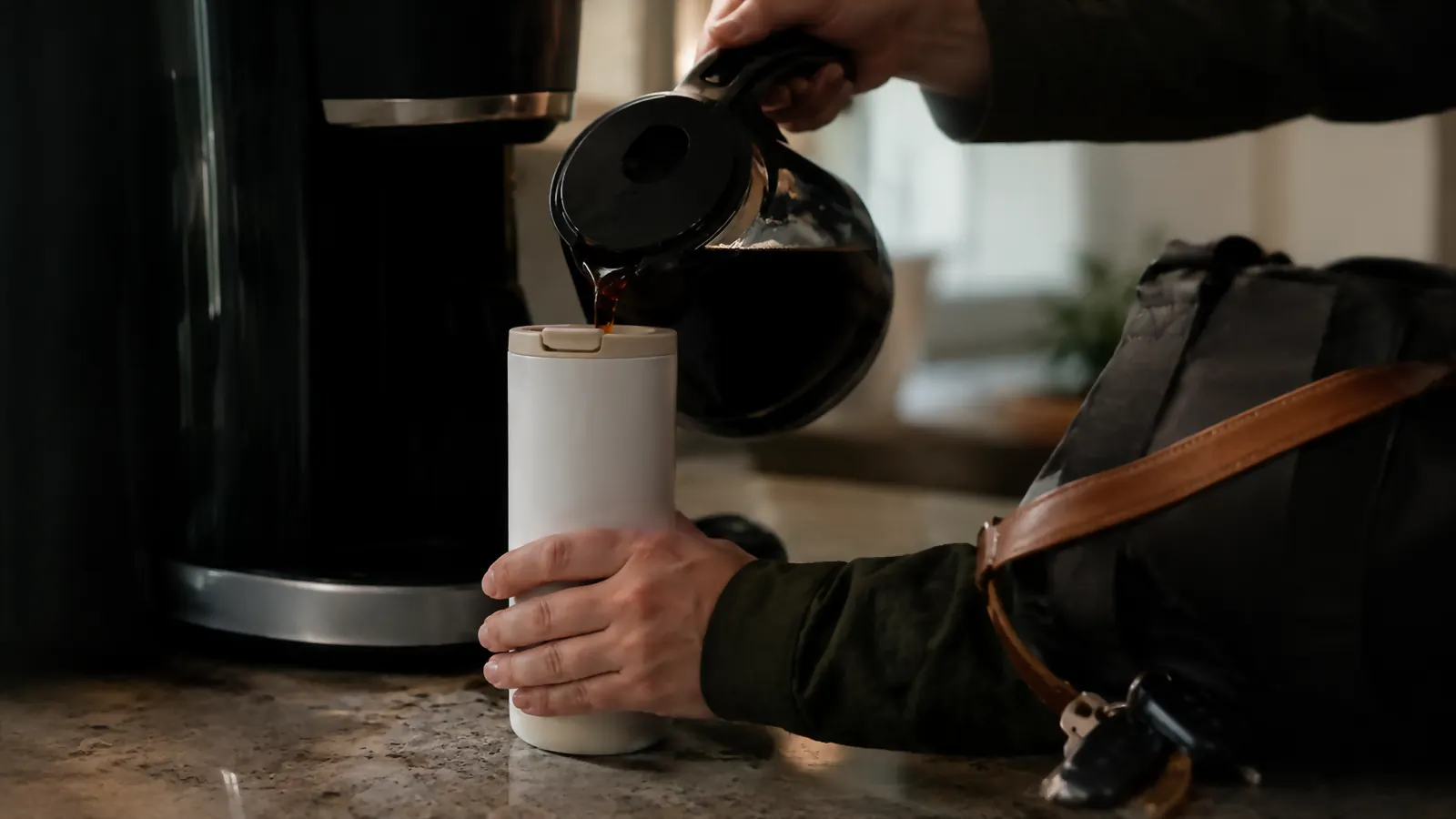 A hand pouring coffee from a small drip machine into the Owala SmoothSip tumbler on a countertop with a travel bag strap visible in frame