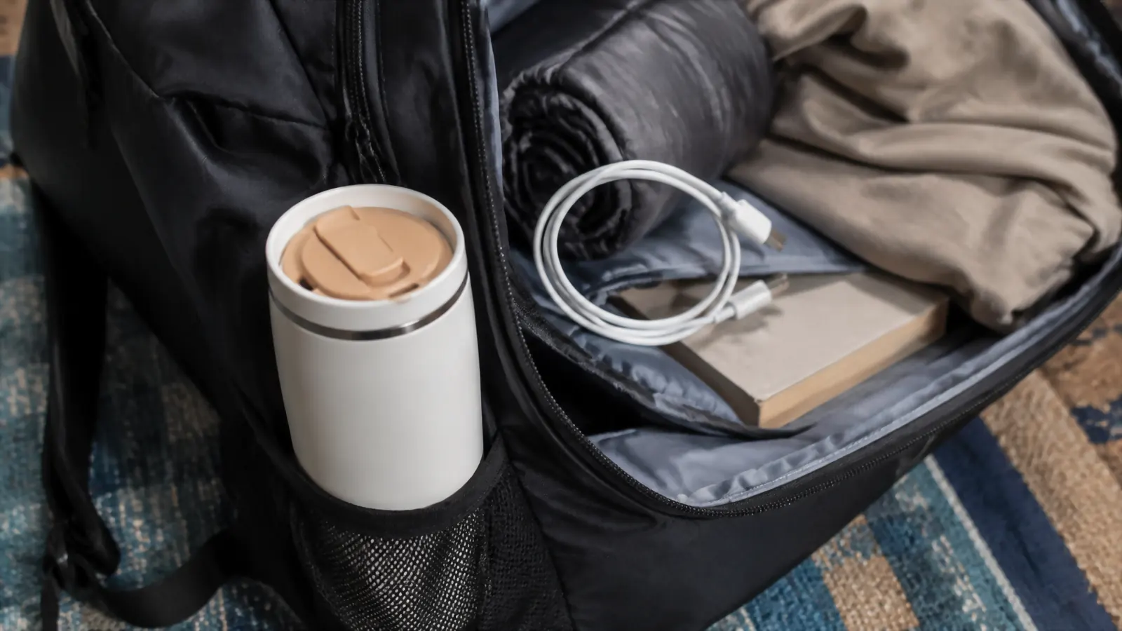 The Owala SmoothSip tumbler with its slider lid open, tucked into the side pocket of an open backpack next to a phone charger